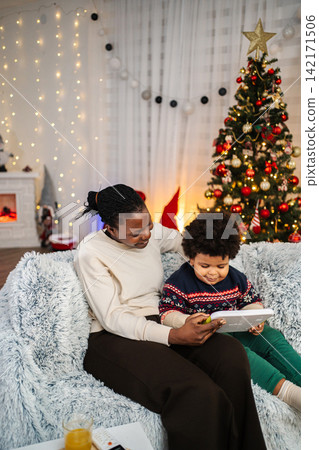 Mother and child enjoying tablet at Christmas with holiday tree Mother and child enjoying tablet at Christmas with holiday tree 142171506