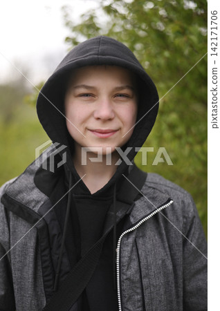 Portrait of young boy wearing black hood outdoors against green foliage background. Portrait of young boy wearing black hood outdoors against green foliage background. 142171706