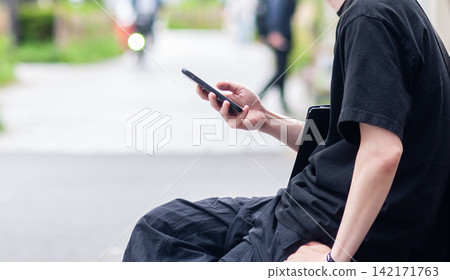 一名年輕男子在街上看著他的智慧型手機。 一名年輕男子在街上看著他的智慧型手機。 142171763