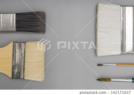 Overhead view of different professional artist paint brushes, large flat wall brushes, and fine detail brushes laid out neatly on a solid grey backdrop with ample copy space for creative design work 142171837