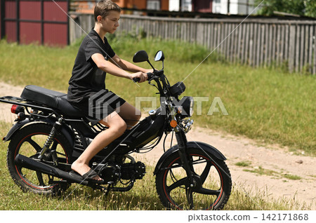 Young boy rides black moped along rural dirt road. Young boy rides black moped along rural dirt road. 142171868
