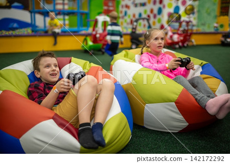 Two children holding controllers and playing video games in bean bag chairs Two children holding controllers and playing video games in bean bag chairs 142172292