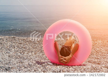 Woman beach inflatable relaxing on a pebble shore framed by a pink swim ring during a peaceful summer sunset Woman beach inflatable relaxing on a pebble shore framed by a pink swim ring during a peaceful summer sunset 142172802