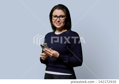 Smiling 40s mature woman in glasses holding smartphone on grey background Smiling 40s mature woman in glasses holding smartphone on grey background 142172910