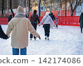 People holding hands while ice skating together on outdoor city winter rink. Natural back view illustrates friendship, togetherness, social recreation, seasonal leisure 142174086