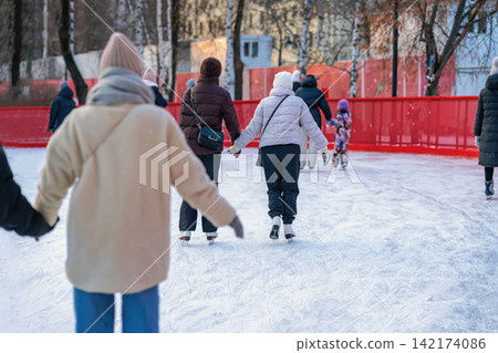 People holding hands while ice skating together on outdoor city winter rink. Natural back view illustrates friendship, togetherness, social recreation, seasonal leisure People holding hands while ice skating together on outdoor city winter rink. Natural back view illustrates friendship, togetherness, social recreation, seasonal leisure 142174086