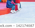 Ice Skaters Resting at Outdoor Rink. Line of teenagers in skates sitting, resting side by side on rink edge of outdoor winter rink, seasonal recreation. Ice area winter background, useful copy space 142174087