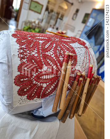 Traditional bobbin lace weaving process with red thread on a pillow using wooden bobbins in a craft setting Traditional bobbin lace weaving process with red thread on a pillow using wooden bobbins in a craft setting 142174213