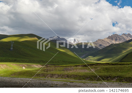 Mountain Landscape with Green Hills, Snowy Peaks, and Dramatic Clouds Mountain Landscape with Green Hills, Snowy Peaks, and Dramatic Clouds 142174695