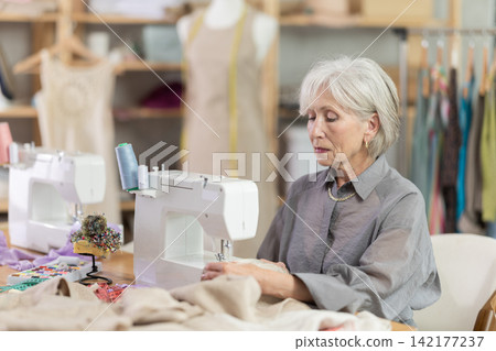 Mature female dressmaker working with sewing-machine in tailor's workshop Mature female dressmaker working with sewing-machine in tailor's workshop 142177237