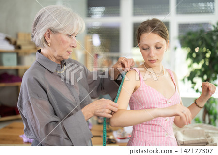 Mature tailoress measuring female client in sewing workshop Mature tailoress measuring female client in sewing workshop 142177307