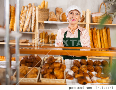 Portrait of elderly woman baker standing behind display case with fresh croissants. Offering to buy fresh baked goods Portrait of elderly woman baker standing behind display case with fresh croissants. Offering to buy fresh baked goods 142177471