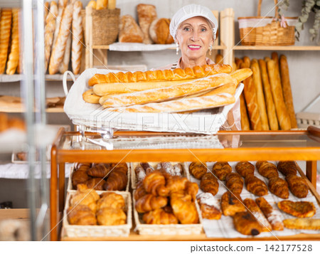 Elderly woman bakery employee offering bread and different baguettes for sale Elderly woman bakery employee offering bread and different baguettes for sale 142177528