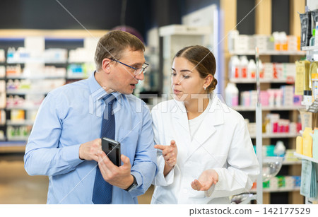 Friendly female pharmacist helping adult man with electronic prescription in phone choosing medical supplies at drugstore Friendly female pharmacist helping adult man with electronic prescription in phone choosing medical supplies at drugstore 142177529
