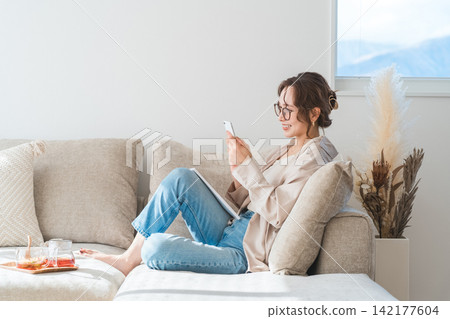 一位亞洲女性坐在客廳的沙發上,看著她的智慧型手機。 一位亞洲女性坐在客廳的沙發上,看著她的智慧型手機。 142177604