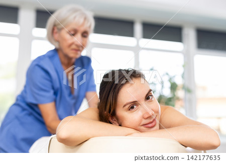 Blurred mature woman actively rubs lower back of young woman during general massage procedure. Blurred mature woman actively rubs lower back of young woman during general massage procedure. 142177635