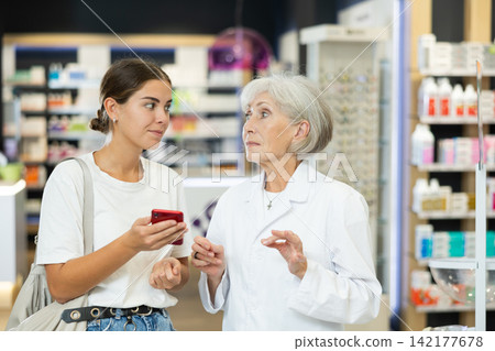 Female shopper shows name of drug on smartphone screen. Female elderly pharmacist helps to find medicine in pharmacy 142177678