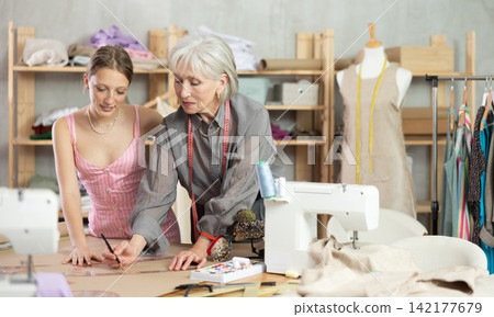Mature tailor draws of a fashion design against the background of a assistant Mature tailor draws of a fashion design against the background of a assistant 142177679