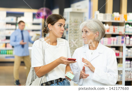 Female shopper shows name of drug on smartphone screen. Female elderly pharmacist helps to find medicine in pharmacy Female shopper shows name of drug on smartphone screen. Female elderly pharmacist helps to find medicine in pharmacy 142177680