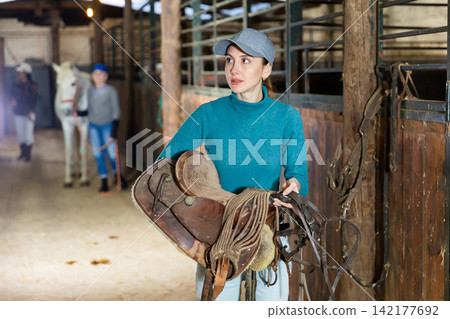 Portrait of an woman in stable with bridle and saddle in her hands 142177692