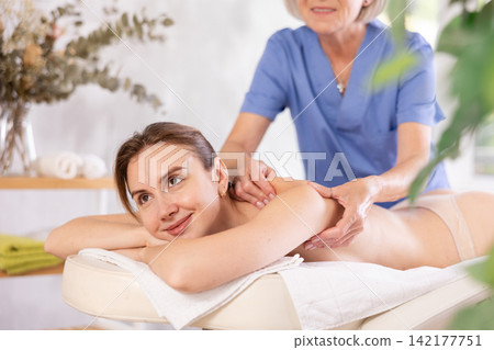 Cropped mature woman actively rubs shoulder of young woman during general massage procedure. Cropped mature woman actively rubs shoulder of young woman during general massage procedure. 142177751