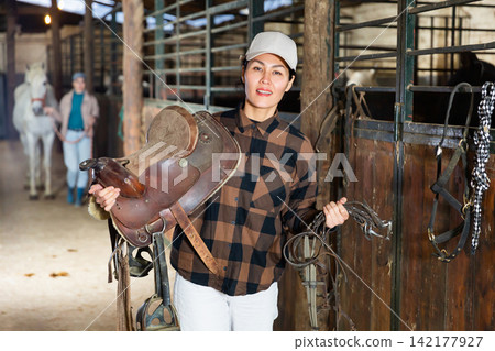 Woman horse breeder carrying leather saddle 142177927