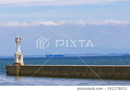 夏日港口景色:蔚藍的大海,防波堤上的白色燈塔,以及地平線上航行的大船。 夏日港口景色:蔚藍的大海,防波堤上的白色燈塔,以及地平線上航行的大船。 142178013