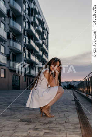 Woman sunset apartment crouching outdoors wearing a white dress near a modern residential building during twilight hours Woman sunset apartment crouching outdoors wearing a white dress near a modern residential building during twilight hours 142180702