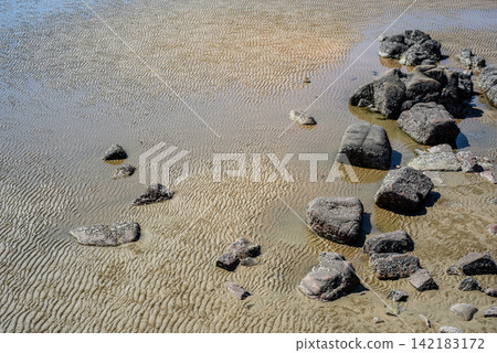 Coastal scene with irregularly shaped dark rocks scattered on a shallow wet shoreline Coastal scene with irregularly shaped dark rocks scattered on a shallow wet shoreline 142183172
