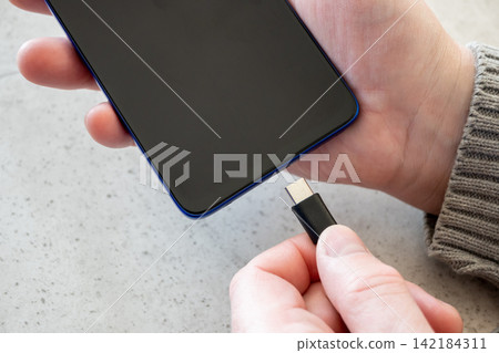 Hand of man girl plugging charger cable into mobile phone on the table. Black cord for smartphone on the stone table. Hand of man girl plugging charger cable into mobile phone on the table. Black cord for smartphone on the stone table. 142184311