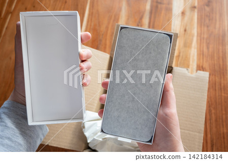 Man unpacking the cardboard box with new mobile phone on the wood table. Smartphone in the hands of a person. Man unpacking the cardboard box with new mobile phone on the wood table. Smartphone in the hands of a person. 142184314