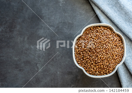 Gray bowl with uncooked roasted buckwheat on the black stone table with napkin for healthy food. Cooking concept. Top view. Space for text. Gray bowl with uncooked roasted buckwheat on the black stone table with napkin for healthy food. Cooking concept. Top view. Space for text. 142184459