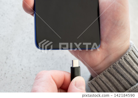 Hand of young girl plugging charger cable into mobile phone on the table. White cord for smartphone. 142184590