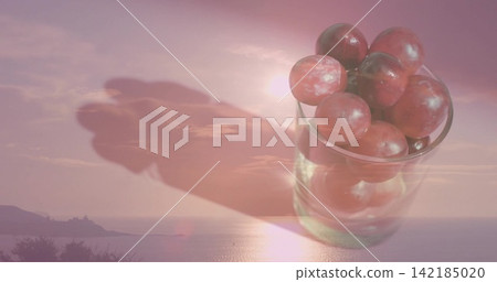 Displaying clear glass holding red grapes over coastal sea, low sun shining, shadow, copy space Displaying clear glass holding red grapes over coastal sea, low sun shining, shadow, copy space 142185020