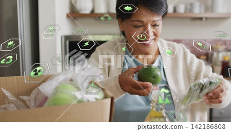 Unpacking senior Indian woman wearing cardigan in kitchen, holding green fruit, box and eco icons 142186808