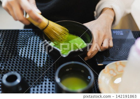 Barista's hand whisking green matcha powder using a traditional bamboo whisk. Barista's hand whisking green matcha powder using a traditional bamboo whisk. 142191863