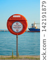Lifebuoy safety station by sea with cargo ship in background. Red lifebuoy safety station with no swimming and no jumping warning signs on a waterfront by calm blue sea, maritime harbor scene 142191879