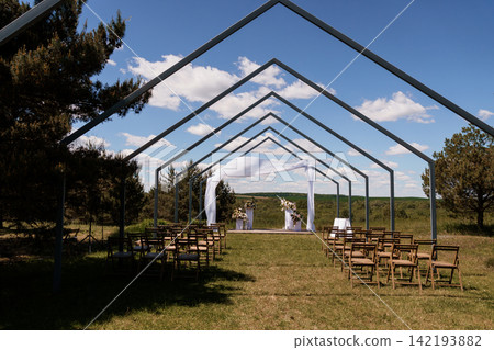 Outdoor wedding ceremony venue with white fabric arch and wooden chairs under metal frame structure in nature. Outdoor wedding ceremony venue with white fabric arch and wooden chairs under metal frame structure in nature. 142193882