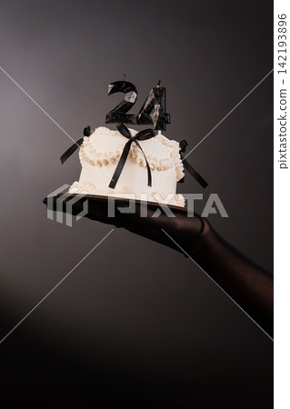 Hand in black glove holding small white cake with black ribbons and number 24 candles on dark grey background. 142193896