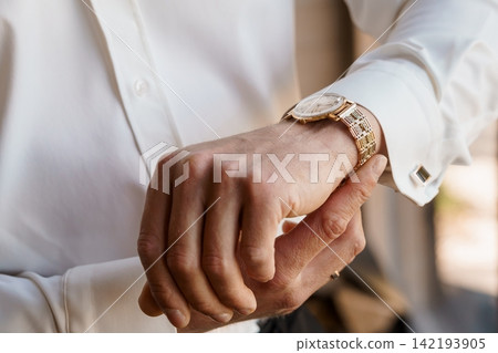 Man in white dress shirt adjusting luxury gold wristwatch with elegant jewelry details in soft lighting. Man in white dress shirt adjusting luxury gold wristwatch with elegant jewelry details in soft lighting. 142193905