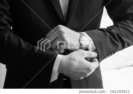 Close up of groom in black tuxedo adjusting white shirt cuff and wearing analog wrist watch in monochrome style. Close up of groom in black tuxedo adjusting white shirt cuff and wearing analog wrist watch in monochrome style. 142193911