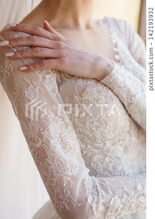 Close up of woman wearing white lace bridal gown with long sleeves and sequins in bright natural light. Close up of woman wearing white lace bridal gown with long sleeves and sequins in bright natural light. 142193932