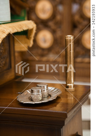 Silver communion cups on a metal tray and brass candle holder on a wooden altar inside a church. Silver communion cups on a metal tray and brass candle holder on a wooden altar inside a church. 142193933