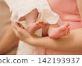 Mother holding newborn baby feet in white lace clothing against a soft pink background. 142193937