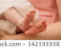 Close up of newborn baby feet resting on adult skin with white lace clothing in soft pink tones. 142193938