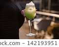 Person holding a chilled green cocktail glass with foam and ice on an outdoor terrace at sunset. 142193951