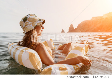 Woman Floating Inflatable Ocean Summer - A woman wearing a hat and sunglasses is floating on an inflatable raft in the ocean, enjoying the summer weather and the view of the cliffs in the distance. Woman Floating Inflatable Ocean Summer - A woman wearing a hat and sunglasses is floating on an inflatable raft in the ocean, enjoying the summer weather and the view of the cliffs in the distance. 142194097