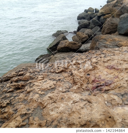 Rugged Coastal Breakwater With Large Brown Boulders Meeting The Gentle Grey Ocean Waves Under A Moody Cloudy Overcast Sky 142194864