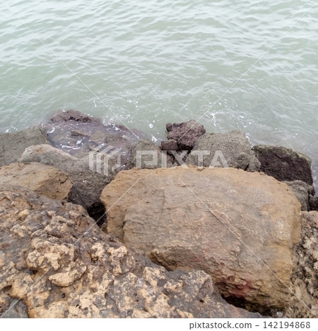 High Angle View Looking Down At Assorted Coastal Boulders And Sea Waves Gently Washing Against The Rocky Shoreline Edge High Angle View Looking Down At Assorted Coastal Boulders And Sea Waves Gently Washing Against The Rocky Shoreline Edge 142194868