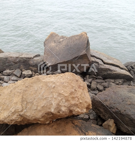 Large Weathered Rocks And Gravel Forming A Coastal Seawall Protecting The Shoreline From The Calm Grey Ocean Water Waves Large Weathered Rocks And Gravel Forming A Coastal Seawall Protecting The Shoreline From The Calm Grey Ocean Water Waves 142194878